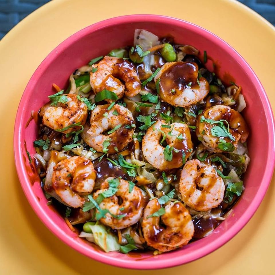 Shrimp and vegetable stir-fry with sauce and cilantro in a pink bowl on a yellow surface.