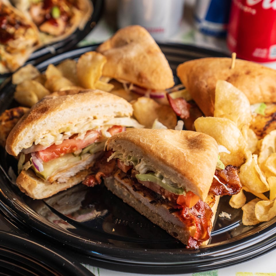 Three sandwiches with chips on a black plate, with tacos and a soda in the background.
