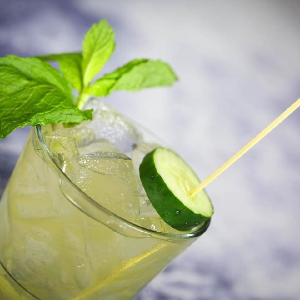 Refreshing cocktail with ice, garnished with fresh mint leaves and a cucumber slice on a skewer.