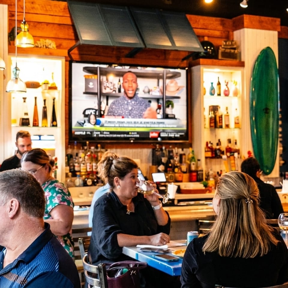People enjoying drinks and conversation at a lively bar with TVs and surfboard decor.