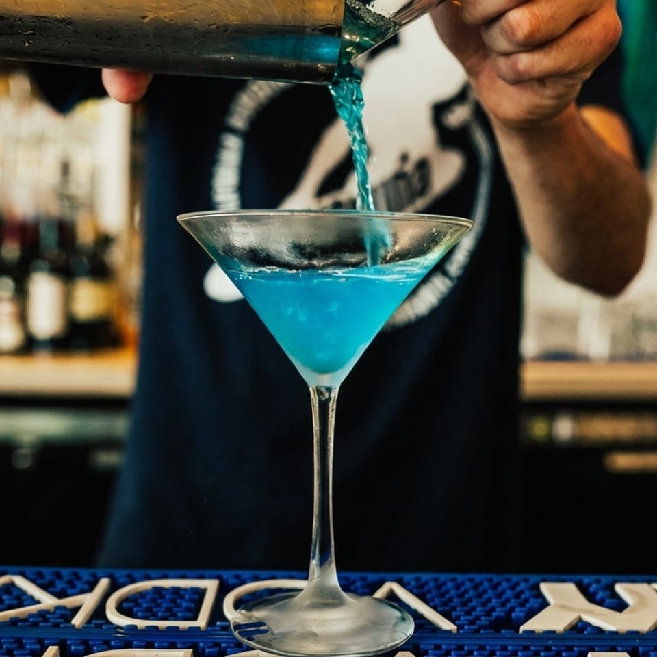 Bartender pouring a blue cocktail from a shaker into a martini glass on a bar mat.