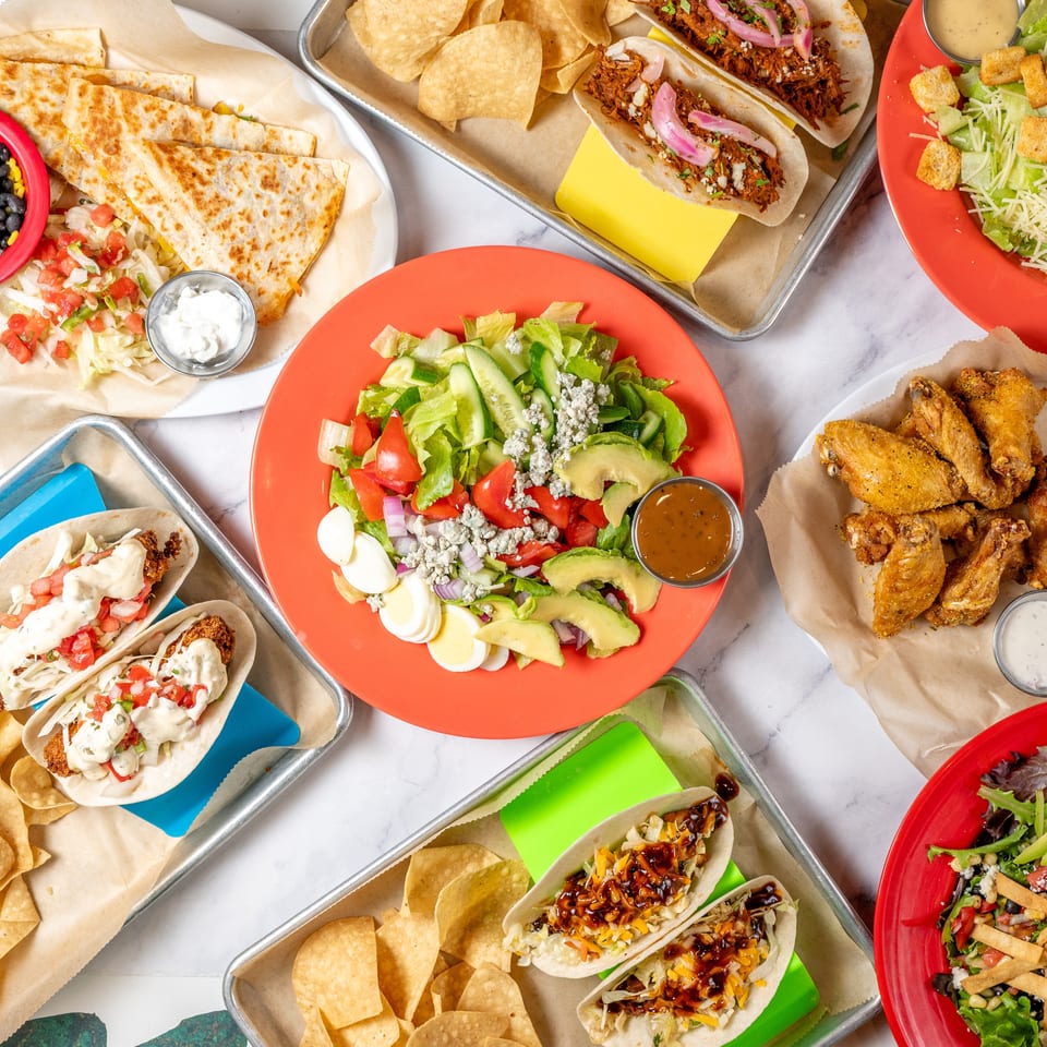 Overhead view of a feast of quesadillas, tacos, salads, chicken wings, and chips on a marble table.