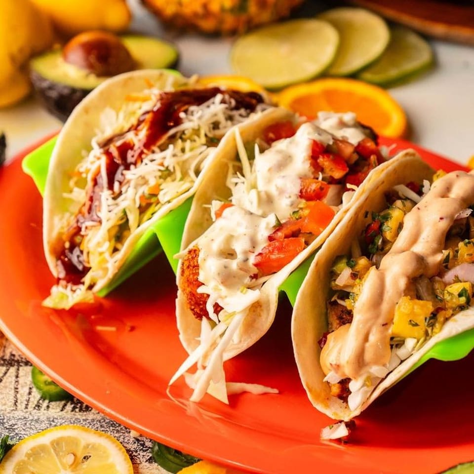Three vibrant tacos with various fillings and sauces, served on an orange plate with citrus slices.