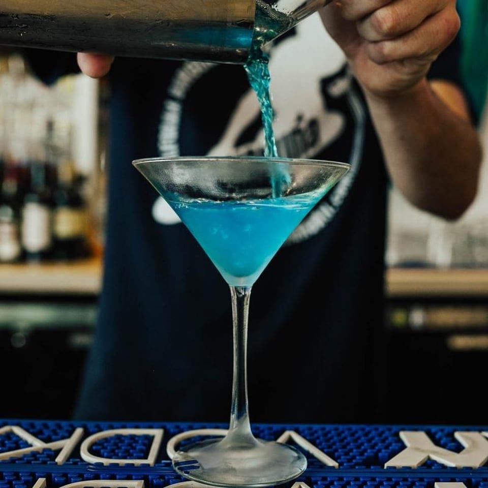 Bartender pouring a vibrant blue cocktail from a shaker into a martini glass.