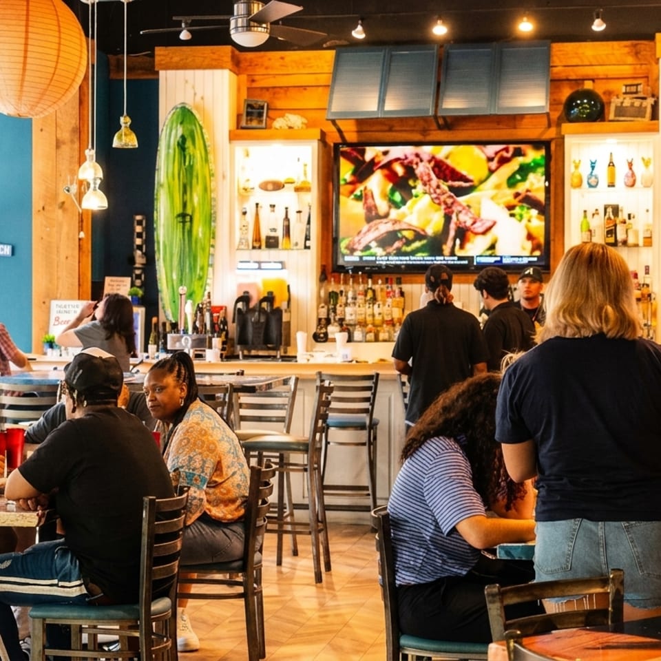 Lively restaurant interior with customers dining, a busy bar, and surf-themed decor.