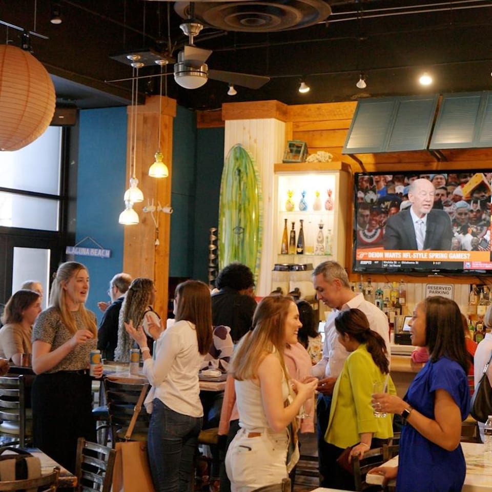People socializing in a lively restaurant bar with a TV, surfboard decor, and hanging lights.