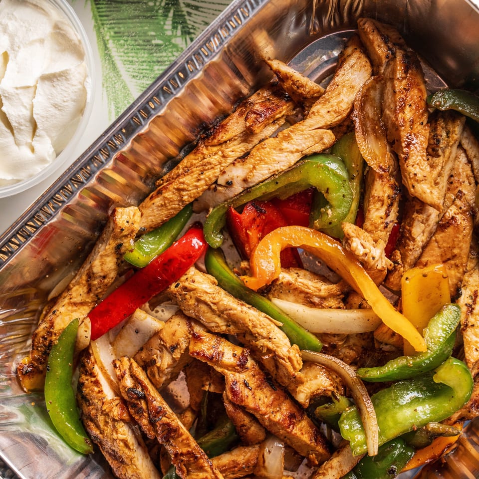 Grilled chicken fajitas with colorful bell peppers and onions in a foil container, with a side of sour cream.