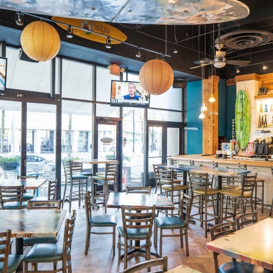 Casual restaurant interior featuring tables, a bar, multiple TVs, and large windows with street views.