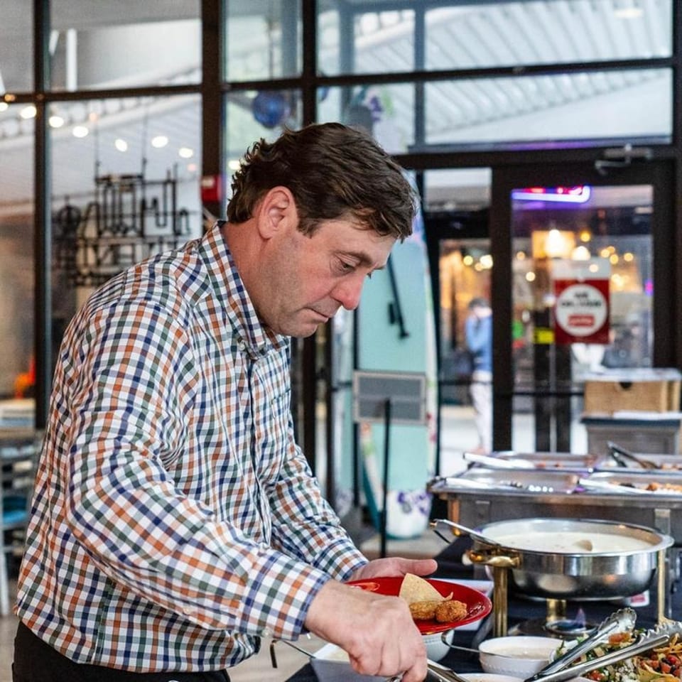 Man in plaid shirt serving himself food from a buffet line with chafing dishes.